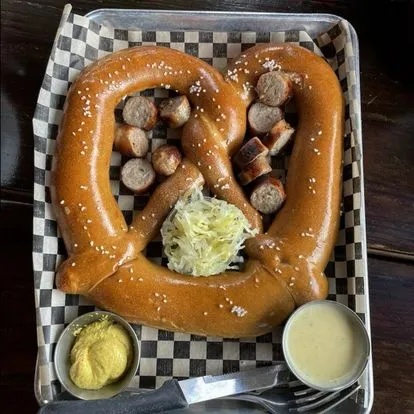 Giant pretzel with sausages, mustard, and cheese sauce on a checkered tray.