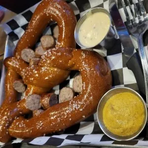 Pretzel with sausage slices, mustard, and cheese dip on a checkered tray.